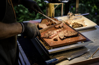 Hand mit schwarzem Handschuh schneidet gegrilltes Brisket auf einem Holzbrett @ 