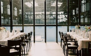 Gedeckte lange Festtafeln in einer Industriehalle mit Glasfront und Blick aufs Wasser @ 