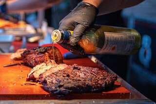 Hand mit schwarzem Handschuh beträufelt aufgeschnittenes gegrilltes Rindfleisch mit Öl @ 