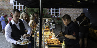Kellner mit Tablett und Grillmeister an offener BBQ-Station vor historischer Backsteinwand @ 